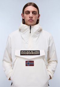 Biała anorakowa kurtka z wysokim kołnierzem, z zamkiem z przodu, naszywką z logo oraz emblematem flagi Norwegii na przedniej kieszeni. Gładka tekstura.