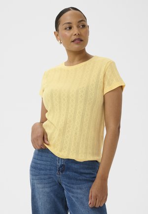 Femme portant un haut à manches courtes jaune à motifs et un jean bleu, posant avec une main dans la poche, regardant sur le côté.