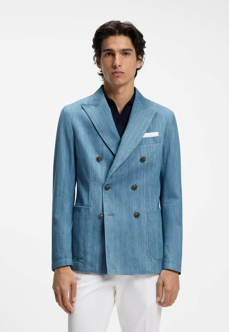 Jeune homme portant un blazer bleu clair croisé avec deux poches avant et un mouchoir blanc, associé à un pantalon blanc.