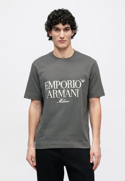Sivé bavlnené tričko s okrúhlym výstrihom a krátkymi rukávmi, s výrazným nápisom "Emporio Armani Milano" v krémovej farbe.