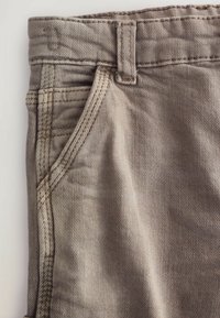 Jean en denim beige avec une vue rapprochée, présentant une poche avant, des passants de ceinture et des détails de couture visibles sur le tissu.