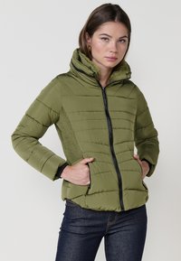 Chaqueta de plumas verde oliva con cierre de cremallera, capucha y bolsillos laterales; presenta una textura acolchada y detalles en negro en la cremallera.