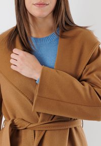 Cappotto marrone avvolgente in tessuto morbido con vita accollata, indossato sopra un maglione a coste blu chiaro. Presenta maniche larghe e dettagli di cucitura diagonale.