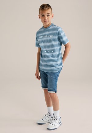 Niebieska koszulka T-shirt tie-dye z napisem "BLUE RIDGE DENIM", zestawiona z podwiniętymi szortami dżinsowymi. Całość uzupełniają białe skarpetki i tenisówki.