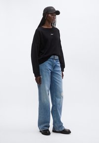 Γυναίκα που φοράει μαύρο καπέλο, μαύρο φούτερ, ανοιχτό μπλε τζιν παντελόνι με φαρδιά πόδια και σκισίματα, και μαύρα παπούτσια, στέκεται μπροστά σε απλό λευκό φόντο.