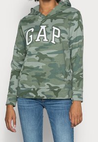 Camo-hoodie in groene tinten met wit "GAP"-logo. Voorzien van een zak aan de voorkant en een capuchon met trekkoord. Gemaakt van zachte, gestructureerde stof.