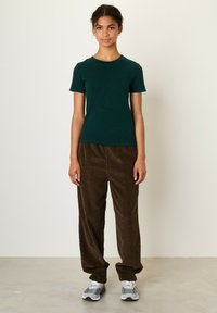 Temno zelena bombažna majica s kratkimi rokavi, v kombinaciji z ohlapnimi rjavimi kordnimi hlačami. Manekenka nosi sivo obutev in stoji na nevtralni podlagi.