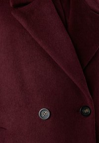 Ingrandimento di un cappotto di lana bordeaux scuro con ampi revers e due bottoni neri a quattro fori.