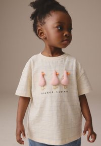 Kremfarvet t-shirt med korte ærmer med tre lyserøde andeapplikationer og teksten "ALWAYS CURIOUS." Designet med et let gittermønster.