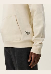 Crème-kleurig sweatshirt met een voork pocket, ribbelmanchetten en een klein zwart geborduurd "LD" logo aan de onderzijde links.