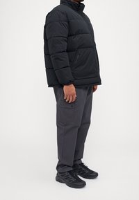 Czarna puchowa kurtka z pikowanym wzorem, wysokim kołnierzem i bocznymi kieszeniami na zamek, zestawiona z szarymi spodniami cargo i czarnymi butami sportowymi.