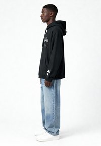 Felpa nera con grafica a croce frontale e testo bianco, abbinata a jeans baggy azzurri e sneakers bianche. Vista laterale, design semplice.