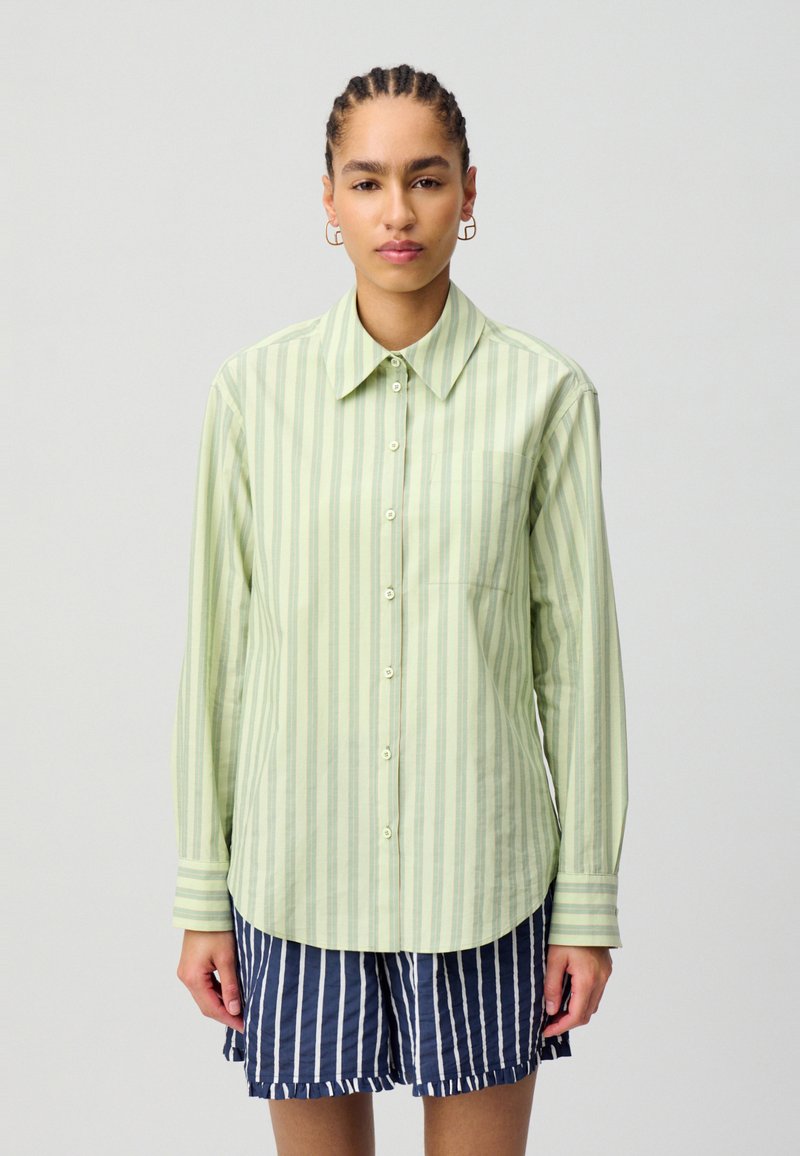 Camicia a maniche lunghe verde chiaro con righe verticali, chiusura con bottoni sul davanti e taschino sul petto. Abbinata a calzoni corti a righe blu e bianche.