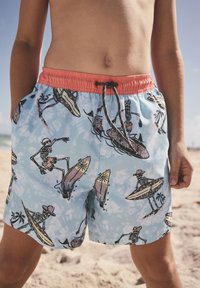 Child wearing blue swim shorts with orange waistband featuring skeletons surfing and skateboarding on a sunny beach.