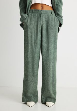 Grønne bukser med vide ben og lommer, båret sammen med matchende blazer og hvide spidse sko, vist fra talje til fødder.