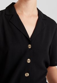 Donna che indossa un blouse nero a maniche corte con colletto a punte e tre grandi bottoni marrone chiaro lungo il davanti.