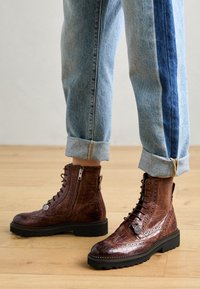 Bottines en cuir marron avec une texture crocodile, design à lacets, fermeture éclair sur le côté et épaisse semelle en caoutchouc noir, associées à un jean bleu clair retroussé.