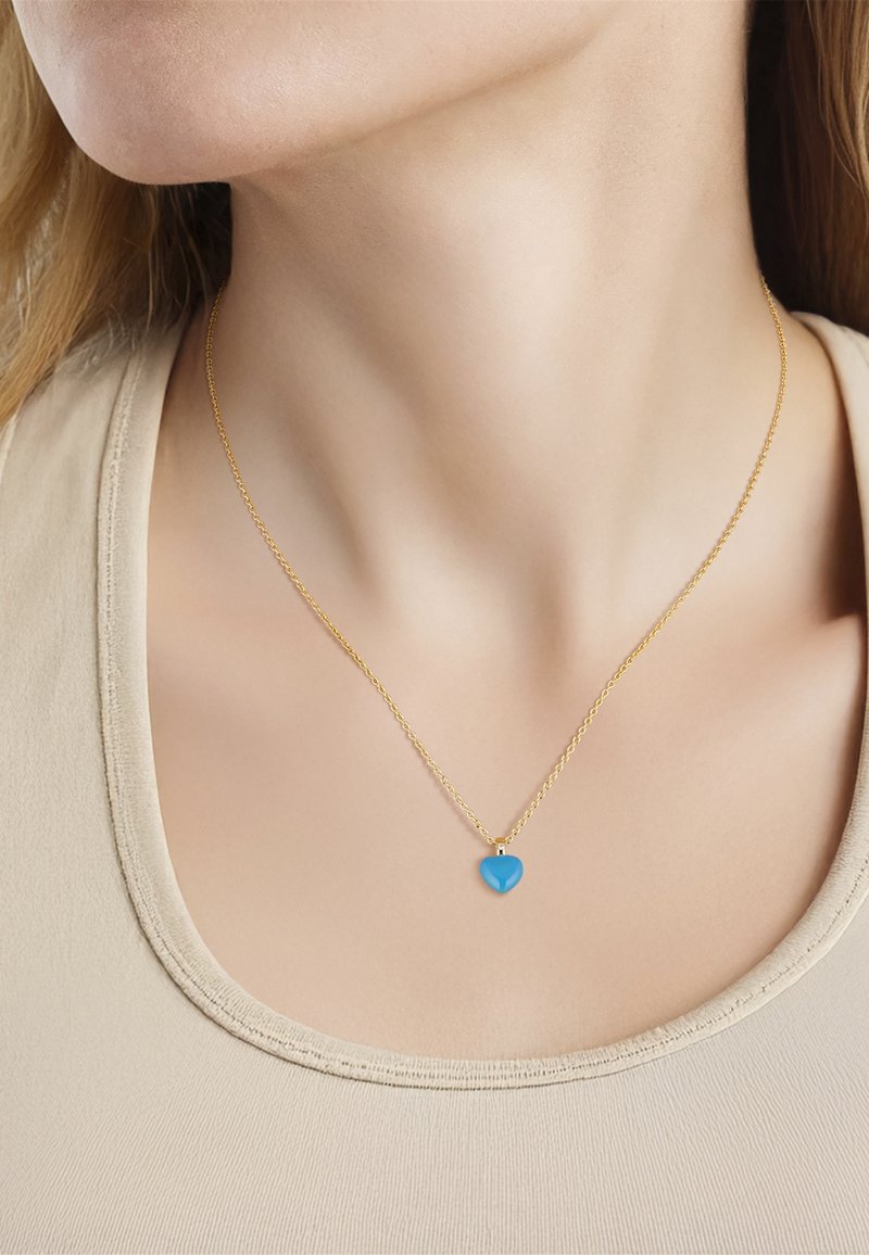Collar de cadena de oro con un pequeño colgante azul brillante en forma de corazón que se lleva en el cuello de una persona sobre una camiseta beige.