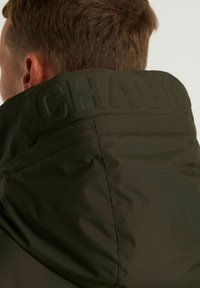 Chaqueta verde oscuro con un cuello alto. Texto grabado "CHASING" en el cuello, textura suave y detalles de costura visibles en las costuras.