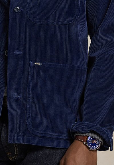 Άνδρας που φοράει μπλε ναυτικό μπουφάν κοτλέ με μεγάλες μπροστινές τσέπες και ένα ρολόι με μπλε καντράν και καφέ δερμάτινο λουρί.