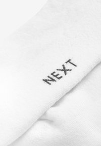 Weißer Baumwollsocken mit einer weichen Textur, der mit einem schwarzen "NEXT"-Logo an der Seite bestickt ist. Die Nahaufnahme hebt die Details des Stoffes hervor.