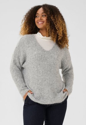 Une femme souriante porte un pull gris ample et duveteux sur un haut blanc et un jean foncé, devant un fond clair uni.