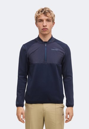Ung mand med kort krøllet blondt hår iført en navyblå Calvin Klein lynlås-pullover og beige bukser, stående mod en ensfarvet baggrund.