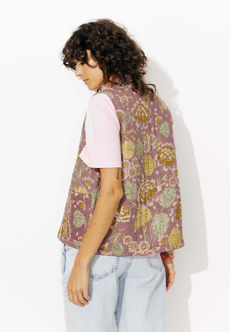 Gilet matelassé fleuri en mauve avec des accents verts et dorés, doté d'une coupe décontractée et de poches latérales, associé à un t-shirt rose pâle.