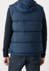 Mörkblå pufferväst med huva, med kviltat mönster och slät yta, bärs över en mörk tröja, i kombination med denimjeans.