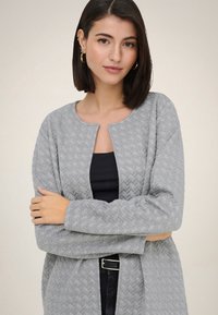 Femme aux cheveux courts et foncés, portant des boucles d'oreilles en anneau dorées et un cardigan gris texturé ouvert sur un haut noir, les bras croisés.