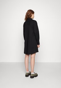 Vestido preto de camisa com mangas longas, corte direito, painéis texturizados e punhos com botões. Usado com calçado verde robusto e meias brancas.