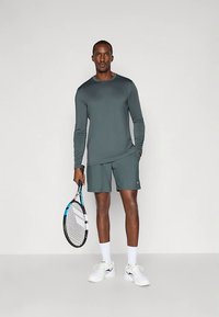 Άνδρας με teal μακρυμάνικη φανέλα και σορτσάκι κρατάει ένα ρακέτα τένις, στέκεται μπροστά σε ένα απλό ανοιχτό γκρι φόντο.