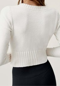 Kobieta z długimi włosami nosi białą, prążkowaną, krótką bluzkę i czarne legginsy, ukazana z tyłu, podkreślająca fason swetra.