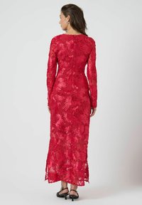 Lang rød aftenkjole med lange ærmer, blomsterbroderi og en tætsiddende silhuet. Har lynlås bagpå og sideslids. Glat tekstur.