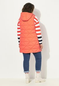 Gilet arancione trapuntato con cappuccio, caratterizzato da un motivo a diamante, abbinato a una camicia a strisce a maniche lunghe e a jeans blu arrotolati, completato da stivali bianchi.