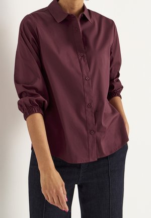 Person iført en bordeauxfarvet skjorte med lange ærmer og elastiske manchetter samt mørke bukser, stående mod en ensfarvet hvid baggrund.