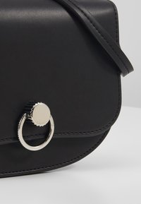 Borsa a tracolla nera in pelle liscia con pattina arrotondata, dotata di un anello in metallo argentato come dettaglio decorativo e cuciture precise.