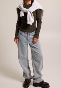 Olivgrön ribbad långärmad topp, ljusblå baggyjeans, grå sweatshirt över axlarna, svarta klumpiga skor med kontrasterande sula.