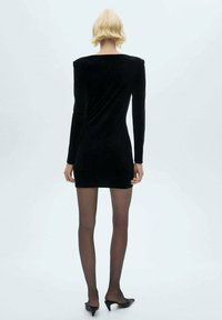 Rochie mini din catifea neagră, cu mâneci lungi, decolteu rotund, design ajustat și textură fină, purtată cu ciorapi negri transparenți și pantofi cu vârful ascuțit.