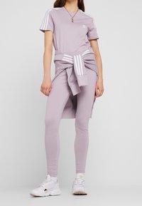 Svetle fialový sportovní outfit zahrnující tričko s bílými pruhy na rukávech a odpovídající legíny; nošeno s bílými teniskami.