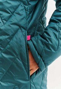 Azurově modrá prošívaná bunda s cik-cak vzorem, boční kapsou a růžovou ozdobnou štítkem. Tkanina má hladký, lesklý povrch.