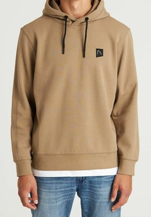 Mann trägt beigen Hoodie mit schwarzen Kordeln, kleinem schwarzem quadratischem Logo auf der Brust, darüber weißes Hemd und blaue Jeans.