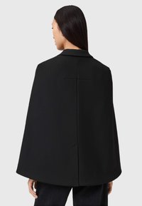 Veste noire en cape avec une silhouette structurée, un col cranté et une texture lisse. Le design au dos présente une couture centrale et aucun dispositif de fermeture visible.