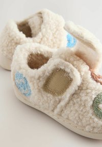 Chaussons en laine beige avec surface texturée, ornés d'accents brodés en bleu, vert et marron. Design sans lacets avec dos ouvert.