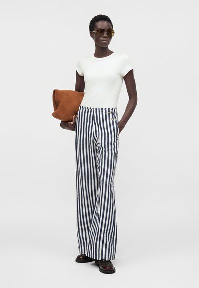 Γυναίκα με λευκό εφαρμοστό T-shirt, μπλε και λευκό κάθετο ριγέ φαρδιά παντελόνια, σκούρα loafers, καφέ oversized clutch και κίτρινα γυαλιά ηλίου aviator.