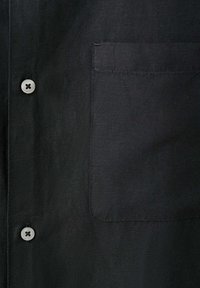 Tissu de chemise noire montrant deux boutons blancs et une seule poche poitrine du côté droit avec des détails de couture visibles.