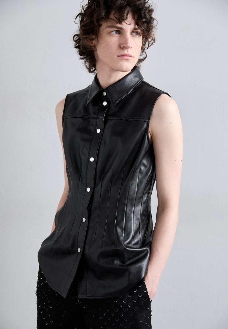 Chemise sans manches en cuir noir avec un col structuré, un devant à boutons et des coutures verticales ; associée à un pantalon noir texturé avec des accents circulaires.