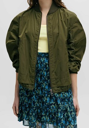 Kobieta w oliwkowo-zielonej kurtce bomberce na jasnżółtym prążkowanym topie i niebieskiej plisowanej spódnicy w kwiaty.