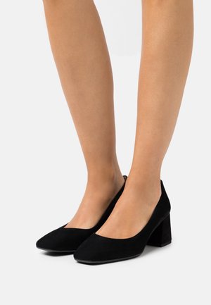 Chaussures noires en daim à talons avec un bout pointu, des côtés bas et un talon block. La texture est douce, offrant une apparence lisse et élégante.