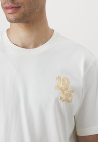 Vitt bomullströja med rund halsringning och korta ärmar. Fram på vänster bröst finns en grafisk text "1956" i gula linjer.